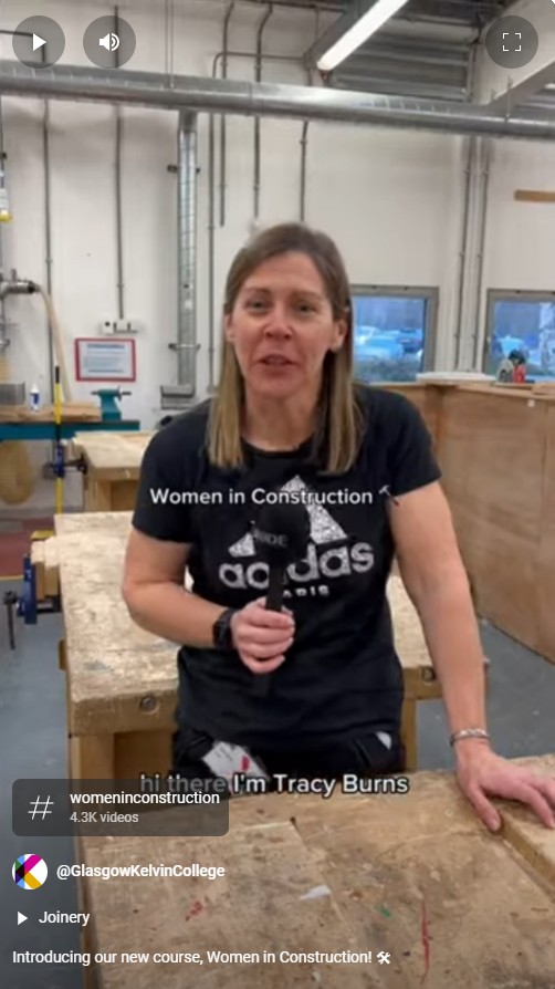 A female lecturer in a black Adidas t-shirt speaking in a joinery workshop at Glasgow Kelvin College, introducing the 'Women in Construction' course, with workbenches and tools visible in the background