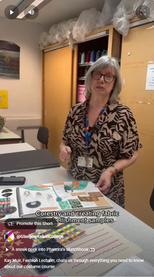 Fashion lecturer explaining costume design techniques, showcasing a sketchbook with fabric embellishment samples in a classroom.