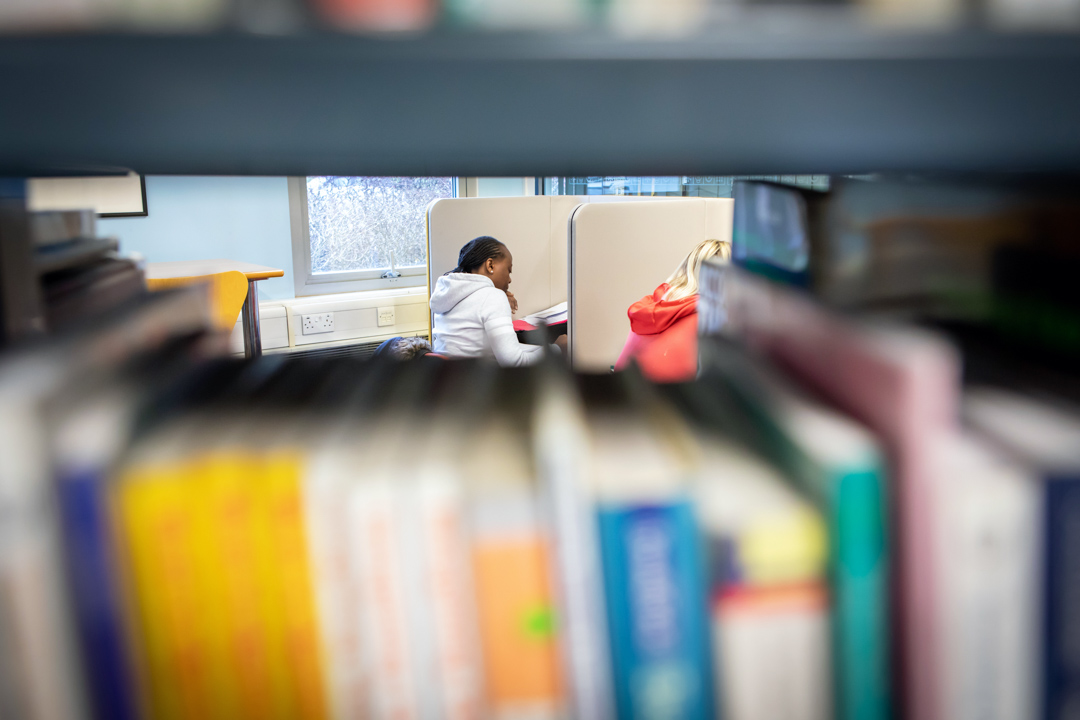 Students studying in individual booths at the East End campus library, viewed through a shelf of books.
