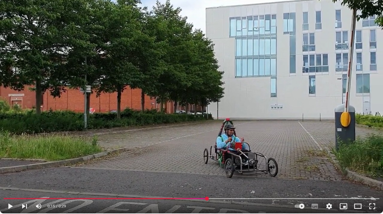 A student in a helmet and blue jacket driving a custom-built electric car in an open parking area outside a modern college building, surrounded by trees