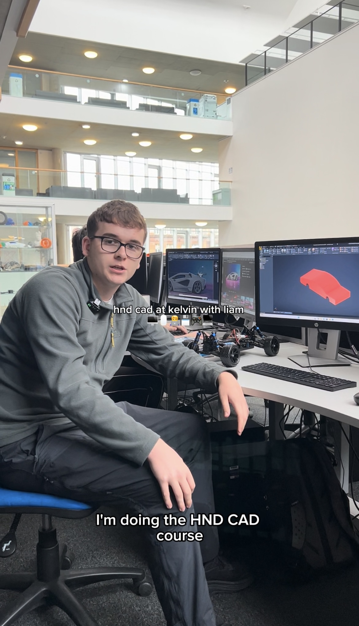 HND CAD student Liam sitting at a computer desk in Glasgow Kelvin College, with 3D car models displayed on dual monitors and a small vehicle prototype on the table.