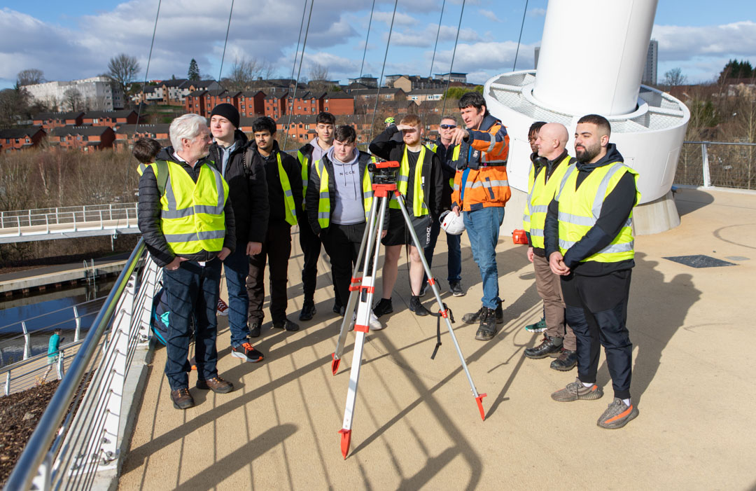 A group of students and lecturers in high-visibility vests gathered around a surveying instrument on the Stockingfield Bridge, listening to an instructor providing guidance.