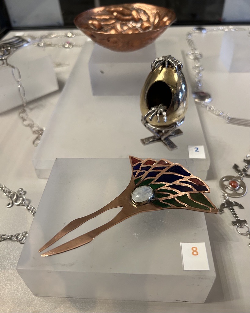 A selection of handcrafted jewellery on display at Craftex 2025, featuring a copper bowl, sculpted egg piece, and an enamelled decorative hairpin.
