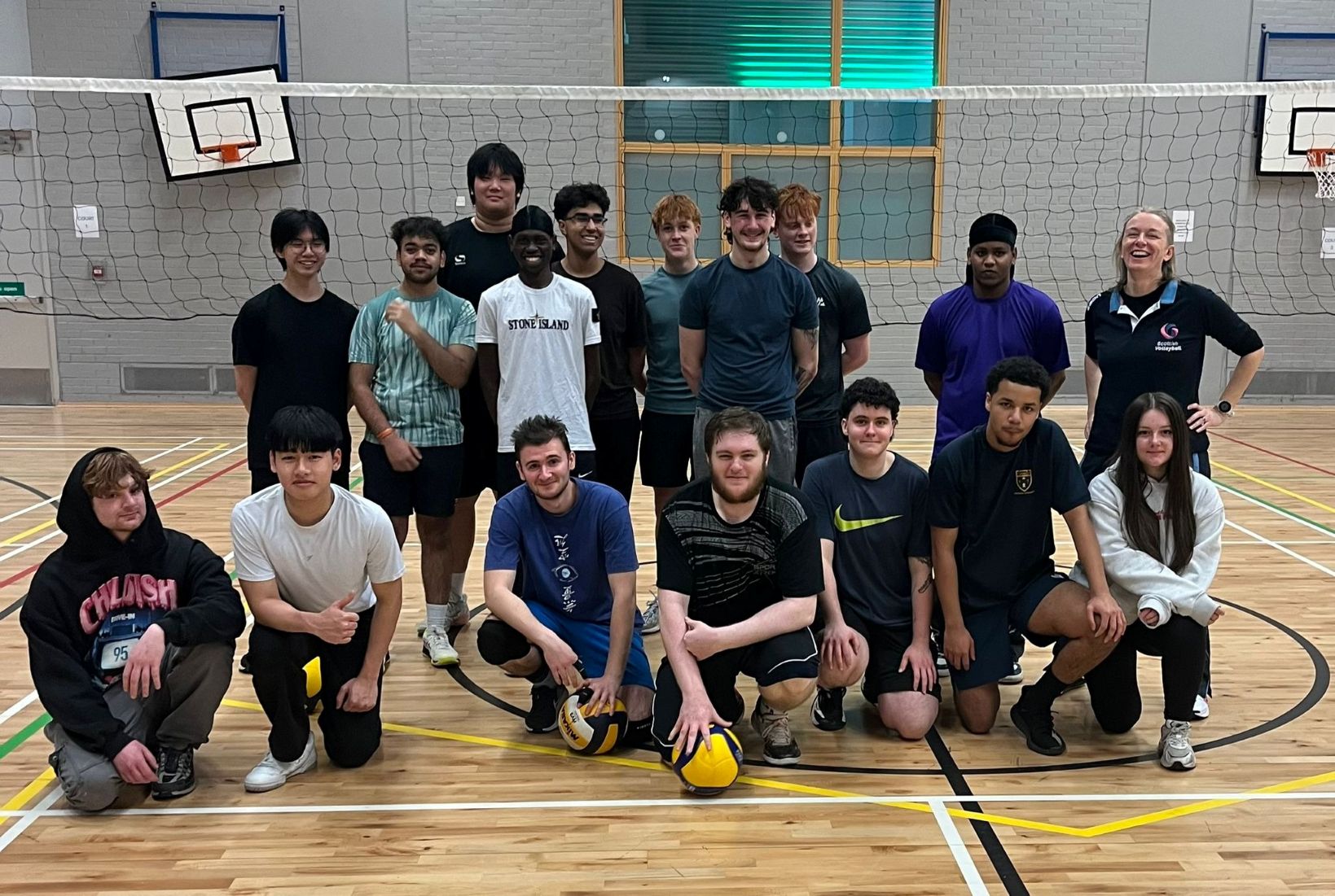 Glasgow Kelvin College students posing as a volleyball team in the sports hall, standing and kneeling in front of the net.
