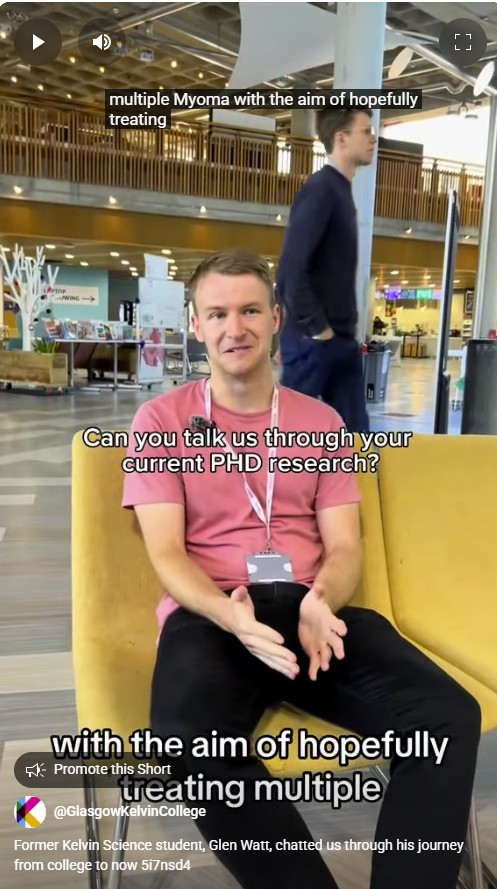 Glen Watt, former Glasgow Kelvin College Science student, sits in the college atrium discussing his PhD research, wearing a pink T-shirt and lanyard.