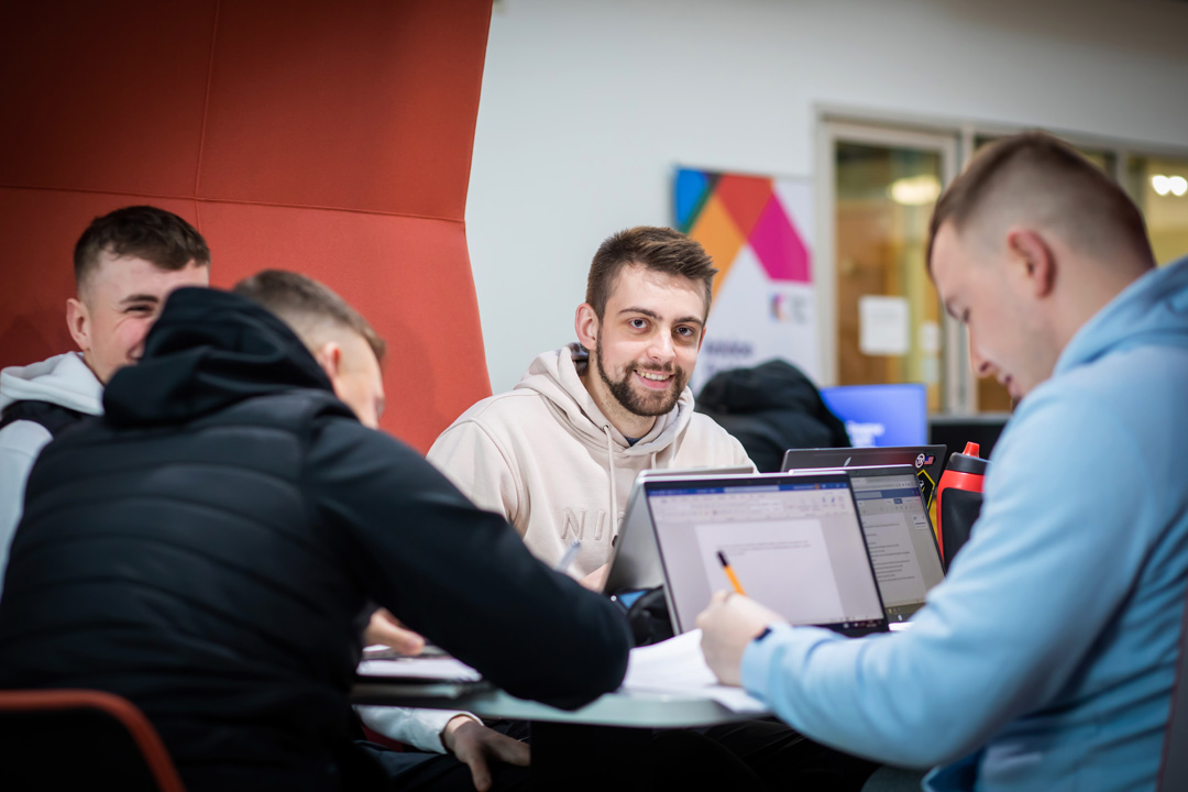 Group of male students working together on laptops and papers in a study pod at Glasgow Kelvin College’s Springburn Campus, with one student smiling at the camera.