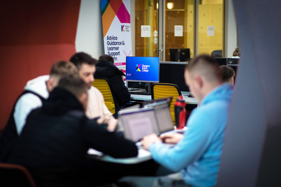Students working together on laptops in a pod area at Glasgow Kelvin College, with a support services banner and desktop monitors in the background.