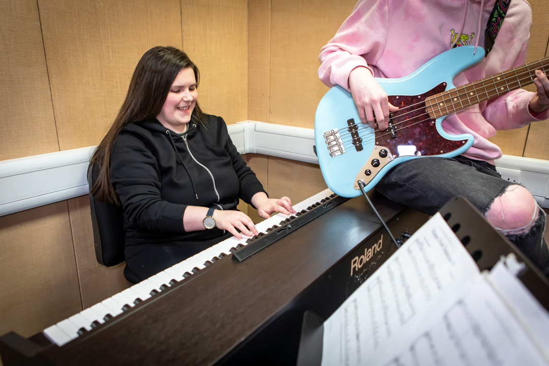 A smiling student in a black hoodie plays a Roland keyboard while another plays a blue bass guitar beside them.