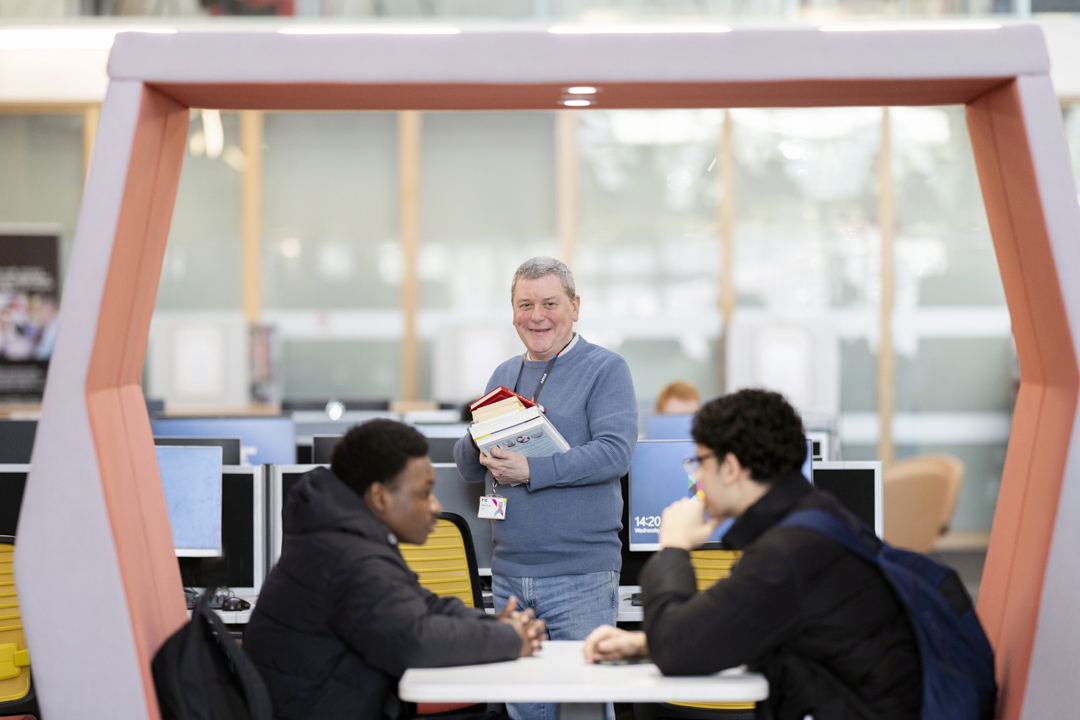 Smiling staff member holding books and standing beside two students in a Glasgow Kelvin College library study pod.
