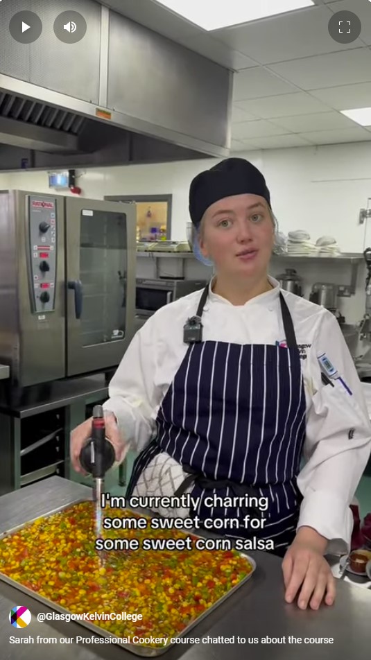 A cookery student in a striped apron and black chef's hat uses a blowtorch to char sweetcorn in a commercial kitchen.