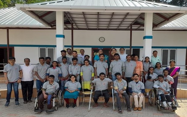 Group photo of staff and youth with disabilities outside a building at IOCL Kerala, with some individuals seated in wheelchairs and others standing, all smiling.