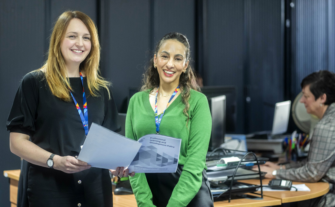 Two Glasgow Kelvin College support staff members smiling and holding documents in an office environment, with a colleague working at a desk in the background.
