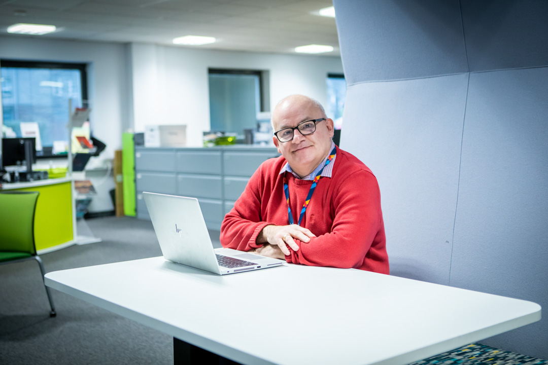 Smiling staff member in a red jumper sitting at a desk with a laptop in a modern office setting at Glasgow Kelvin College