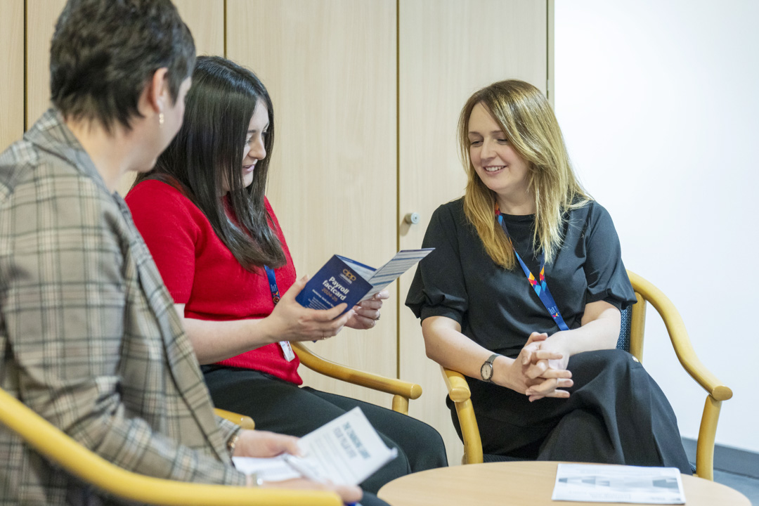 Three Glasgow Kelvin College staff members seated in a meeting room, smiling and discussing printed materials during a relaxed conversation.
