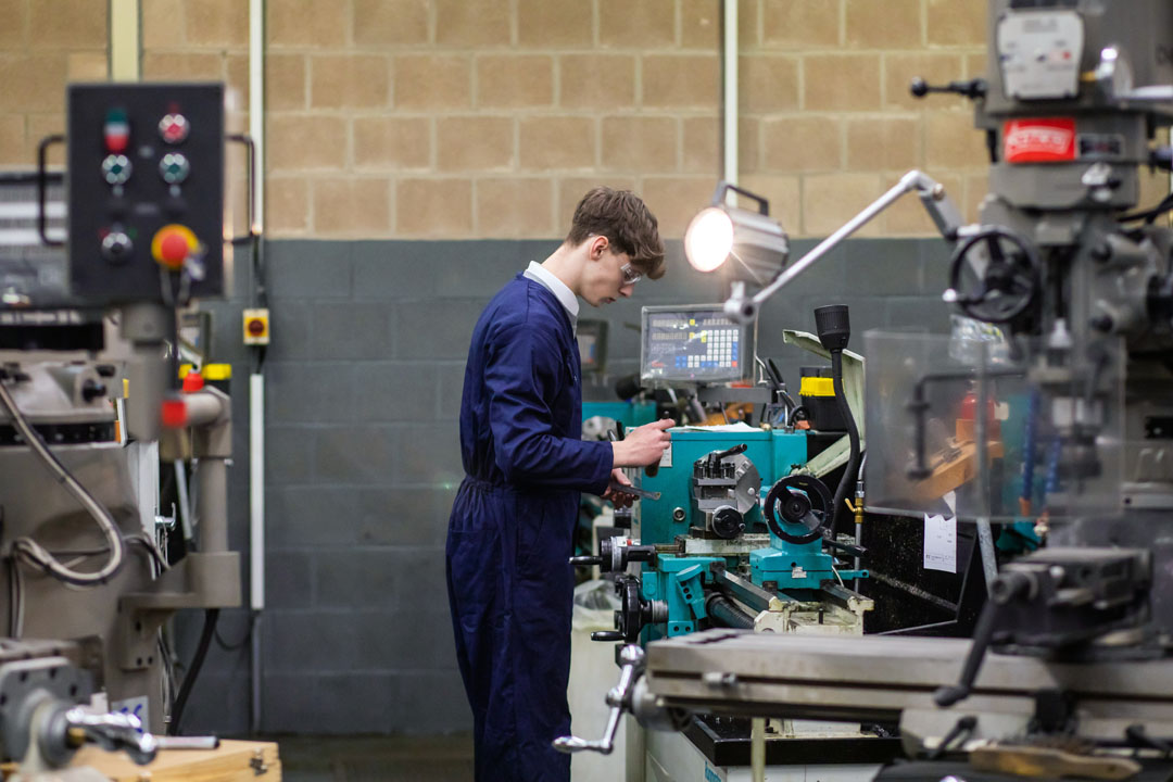 A student in navy-blue overalls operating a lathe machine in a mechanical engineering workshop, focused on precision machining.