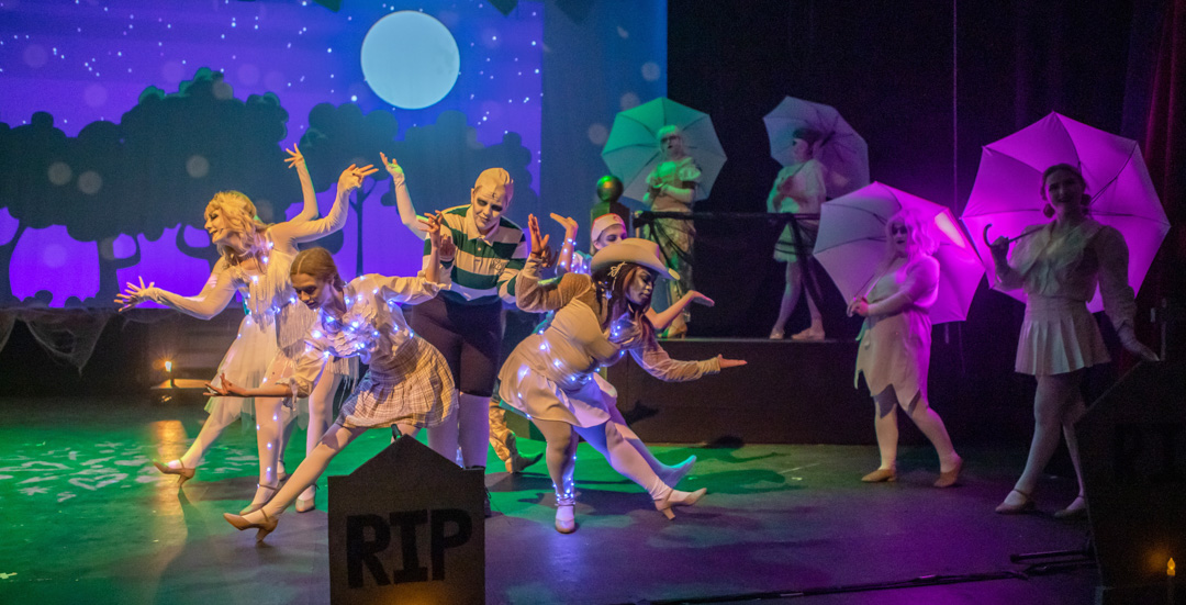 Musical theatre students performing a stylised ensemble dance on stage, with umbrellas, illuminated costumes, and a moonlit backdrop.